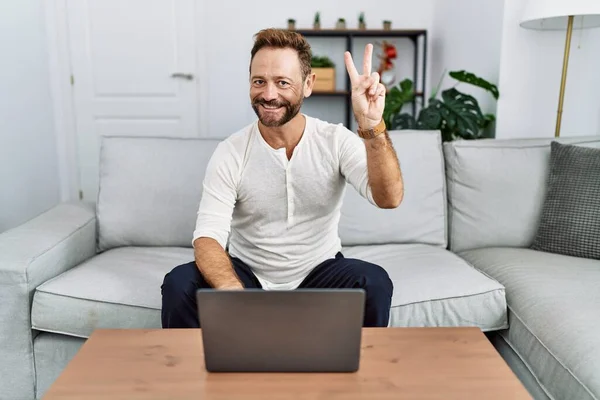 家でノートパソコンを使っている中年の男性は カメラで笑顔で笑顔で勝利のサインを指で行います — ストック写真