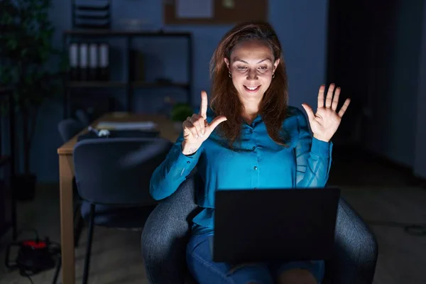 Brunette woman working at the office at night showing and pointing up with fingers number seven while smiling confident and happy.