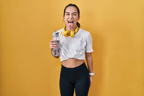 Young south asian woman wearing sportswear drinking water sticking tongue out happy with funny expression. emotion concept.