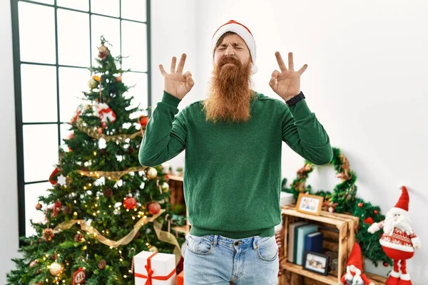 長い髭を生やした赤毛の男がクリスマスツリーでクリスマスの帽子をかぶってリラックスして笑顔で目を閉じて指で瞑想のジェスチャーをしています ヨガのコンセプト — ストック写真