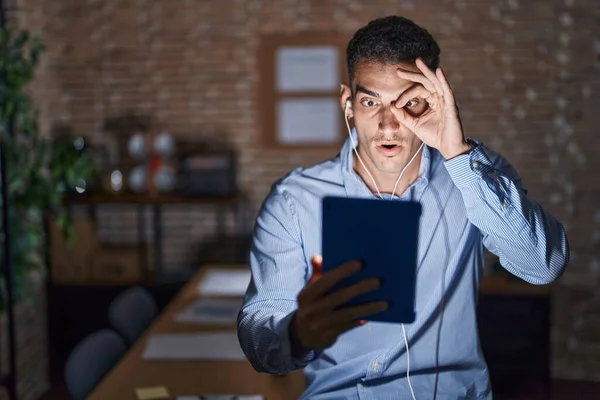 Handsome hispanic man working at the office at night doing ok gesture shocked with surprised face, eye looking through fingers. unbelieving expression.