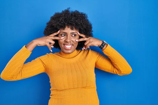 Black woman with curly hair standing over blue background doing peace symbol with fingers over face, smiling cheerful showing victory