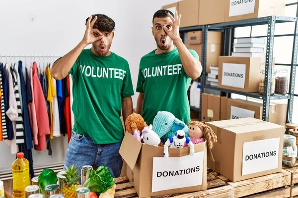 Young gay couple wearing volunteer t shirt at donations stand doing ok gesture shocked with surprised face, eye looking through fingers. unbelieving expression.