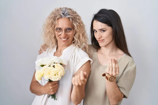 Mother and daughter holding bouquet of white flowers doing money gesture with hands, asking for salary payment, millionaire business