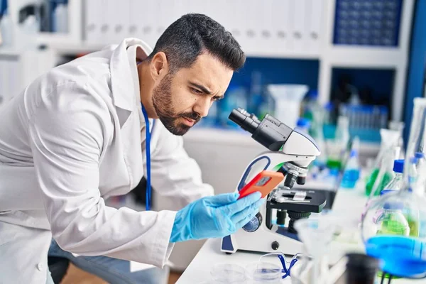 Young Hispanic Man Scientist Using Microscope Smartphone Laboratory — Stock fotografie