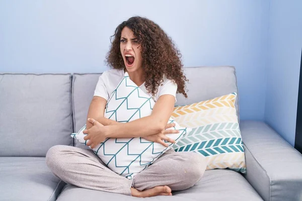 Hispanic woman with curly hair sitting on the sofa at home angry and mad screaming frustrated and furious, shouting with anger. rage and aggressive concept.