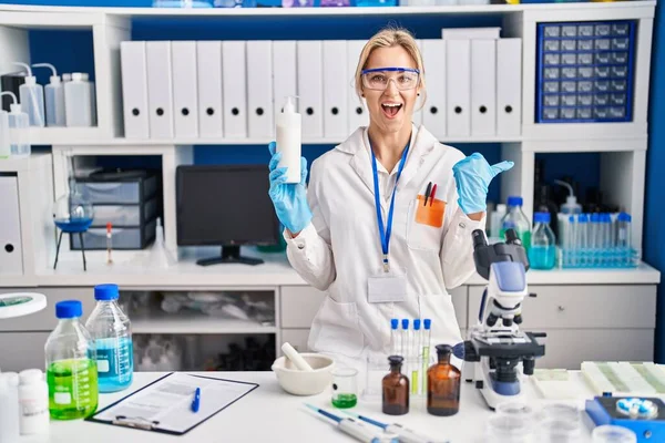 Young caucasian woman working at scientist laboratory holding body lotion pointing thumb up to the side smiling happy with open mouth