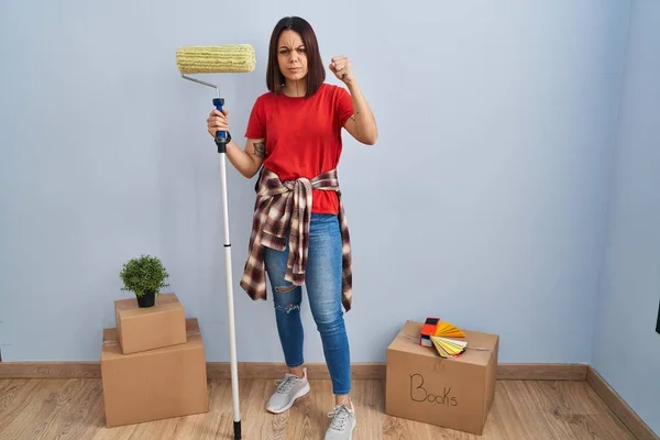 Young hispanic woman painting home walls with paint roller angry and mad raising fist frustrated and furious while shouting with anger. rage and aggressive concept.