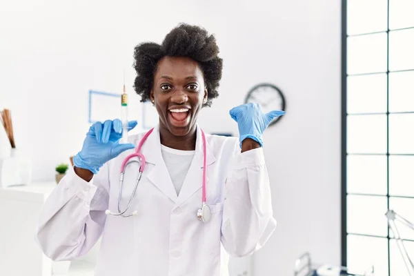 African doctor woman holding syringe at medical clinic pointing thumb up to the side smiling happy with open mouth