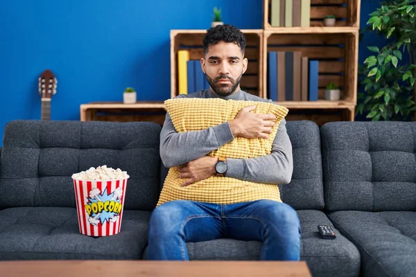 Hispanic man with beard eating popcorn watching a movie at home relaxed with serious expression on face. simple and natural looking at the camera.