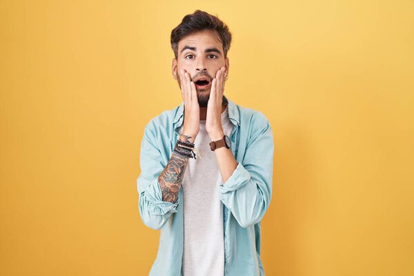 Young hispanic man with tattoos standing over yellow background afraid and shocked, surprise and amazed expression with hands on face 