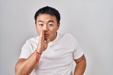 Young chinese man standing over white background hand on mouth telling secret rumor, whispering malicious talk conversation 