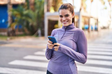 Young beautiful hispanic woman wearing sportswear using smartphone at street