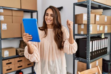 Young hispanic woman working at small business ecommerce using tablet screaming proud, celebrating victory and success very excited with raised arm 