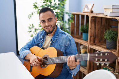 Young hispanic man playing classical guitar sitting on table at home