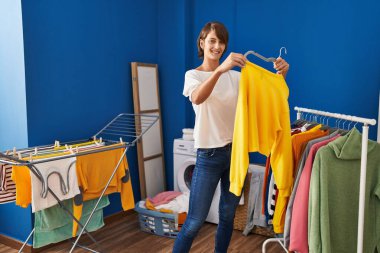 Young beautiful hispanic woman smiling confident holding sweater on clothes rack at laundry room