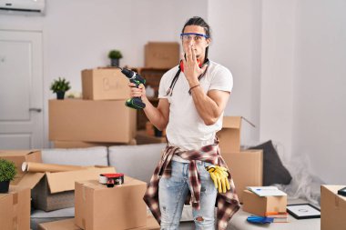 Hispanic man with long hair holding screwdriver at new home covering mouth with hand, shocked and afraid for mistake. surprised expression 
