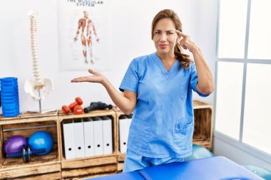 Middle age hispanic physiotherapist woman working at pain recovery clinic confused and annoyed with open palm showing copy space and pointing finger to forehead. think about it. 