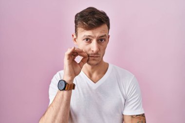 Caucasian man standing over pink background mouth and lips shut as zip with fingers. secret and silent, taboo talking 