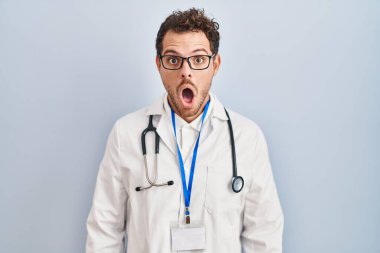 Young hispanic man wearing doctor uniform and stethoscope afraid and shocked with surprise expression, fear and excited face. 