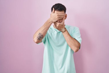 Pembe arka planda duran yakışıklı İspanyol adam gözlerini ve ağzını elleriyle kapatıyor, şaşırmış ve şok olmuş. duygu gizleme 