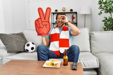 Yakışıklı orta yaşlı adam futbol holiganı evde tezahürat yapıyor gülümsüyor mutlu mesut gülümsüyor tamam el ele imzala parmaklarının arasından bakıyor 