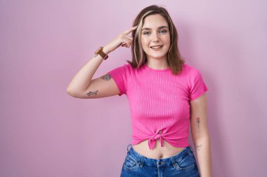 Pembe arka planda duran sarışın beyaz kadın tek parmakla kafaya doğru gülümsüyor, harika bir fikir ya da düşünce, iyi bir hafıza. 
