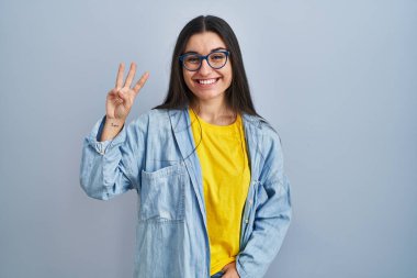 Mavi arka planda dikilen genç İspanyol kadın üç numaralı parmaklarıyla gülümseyerek kendinden emin ve mutlu bir şekilde işaret ediyor.. 