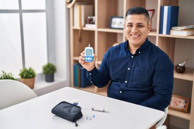 İspanyol genç adam elinde glukometre cihazı tutuyor. Pozitif ve mutlu görünüyor. Ve gülümsüyor. 