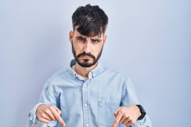 Mavi arka planda dikilen sakallı genç, üzgün ve üzgün görünüyor, parmaklarıyla yönü gösteriyor, mutsuz ve depresif.. 
