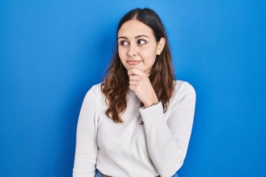 Mavi arka planda dikilen genç İspanyol kadın bir soru hakkında endişeli ve gergin bir şekilde düşünüyor. 