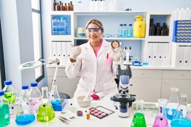 Young hispanic woman working at scientist laboratory doing make up smiling happy and positive, thumb up doing excellent and approval sign 