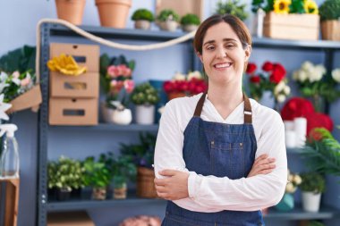 Çiçekçi dükkanında kollarını kavuşturup gülümseyen genç bir kadın.