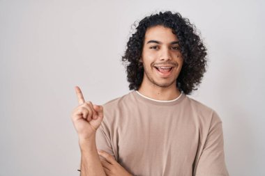 Beyaz arka planda duran kıvırcık saçlı, yüzünde kocaman bir gülümseme olan, eli kameraya bakan İspanyol bir adam.. 