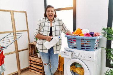 Middle age hispanic woman doing laundry holding detergent bottle celebrating crazy and amazed for success with open eyes screaming excited. 