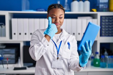 African american woman with braids working at scientist laboratory doing video call with tablet smiling happy and positive, thumb up doing excellent and approval sign 