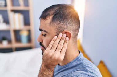 Young hispanic man suffering for ear pain sitting on bed at bedroom