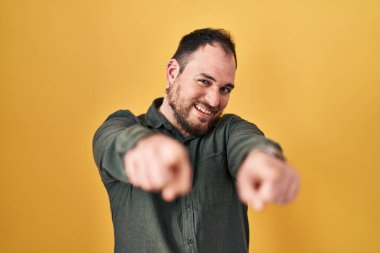 Plus size hispanic man with beard standing over yellow background pointing to you and the camera with fingers, smiling positive and cheerful 