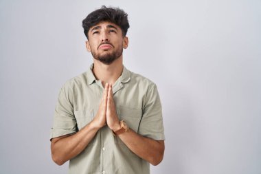Arab man with beard standing over white background begging and praying with hands together with hope expression on face very emotional and worried. begging. 