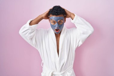 Young hispanic man wearing beauty face mask and bath robe crazy and scared with hands on head, afraid and surprised of shock with open mouth 