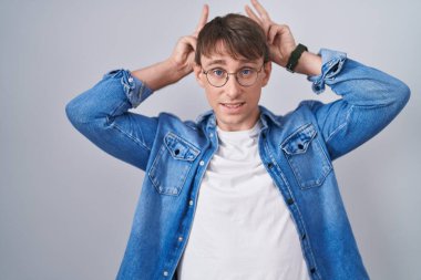 Caucasian blond man standing wearing glasses posing funny and crazy with fingers on head as bunny ears, smiling cheerful 
