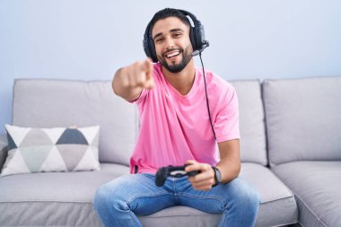 İspanyol genç adam video oyunu oynuyor kanepede oturmuş parmaklarıyla seni ve kamerayı gösteriyor, neşeli ve pozitif gülümsüyor 