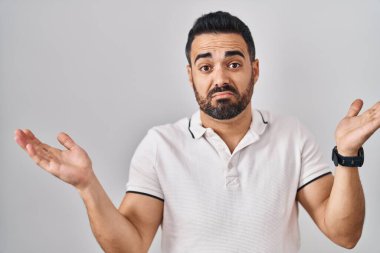 Beyaz arka plan üzerine gündelik kıyafetler giyen sakallı genç İspanyol bir adam. Kolları ve elleri havada, kafası karışmış bir ifade. şüphe kavramı. 