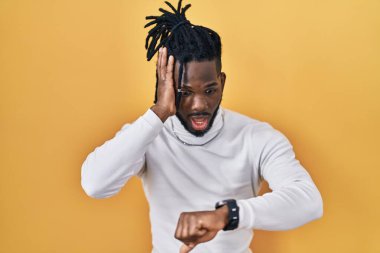 Sarı arka plan üzerine balıkçı yaka kazak giyen Afrikalı bir adam, saati endişeli, geç kalmaktan korkan saatlere bakıyor. 