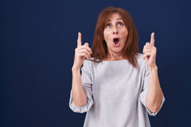 Middle age woman standing over blue background amazed and surprised looking up and pointing with fingers and raised arms. 