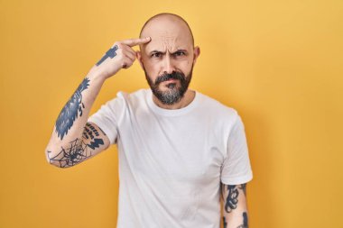 Sarı arka planda duran İspanyol dövmeli genç adam alnındaki sivilceden mutsuz olduğunu gösteriyor, siyah noktalı çirkin bir enfeksiyon. sivilce ve deri problemi 