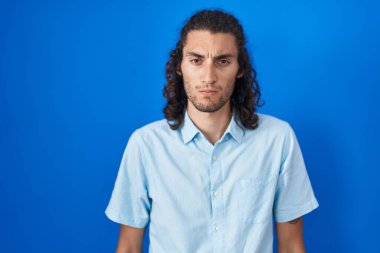 Mavi arka planda duran Latin asıllı genç adam şüpheci ve gergin, sorun yüzünden surat asıyor. negatif kişi. 