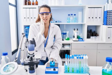 Young hispanic woman working at scientist laboratory touching mouth with hand with painful expression because of toothache or dental illness on teeth. dentist 