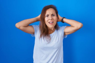 Brunette woman standing over blue background crazy and scared with hands on head, afraid and surprised of shock with open mouth 