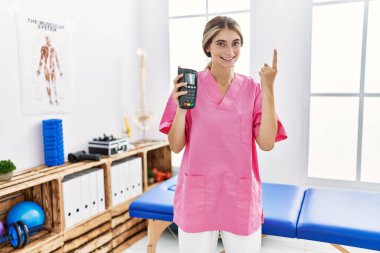 Ağrı kesici kliniğinde çalışan genç fizyoterapist kadın elinde bir fikir ya da soru işareti ile gülümseyen bir veri telefonu tutuyor. 